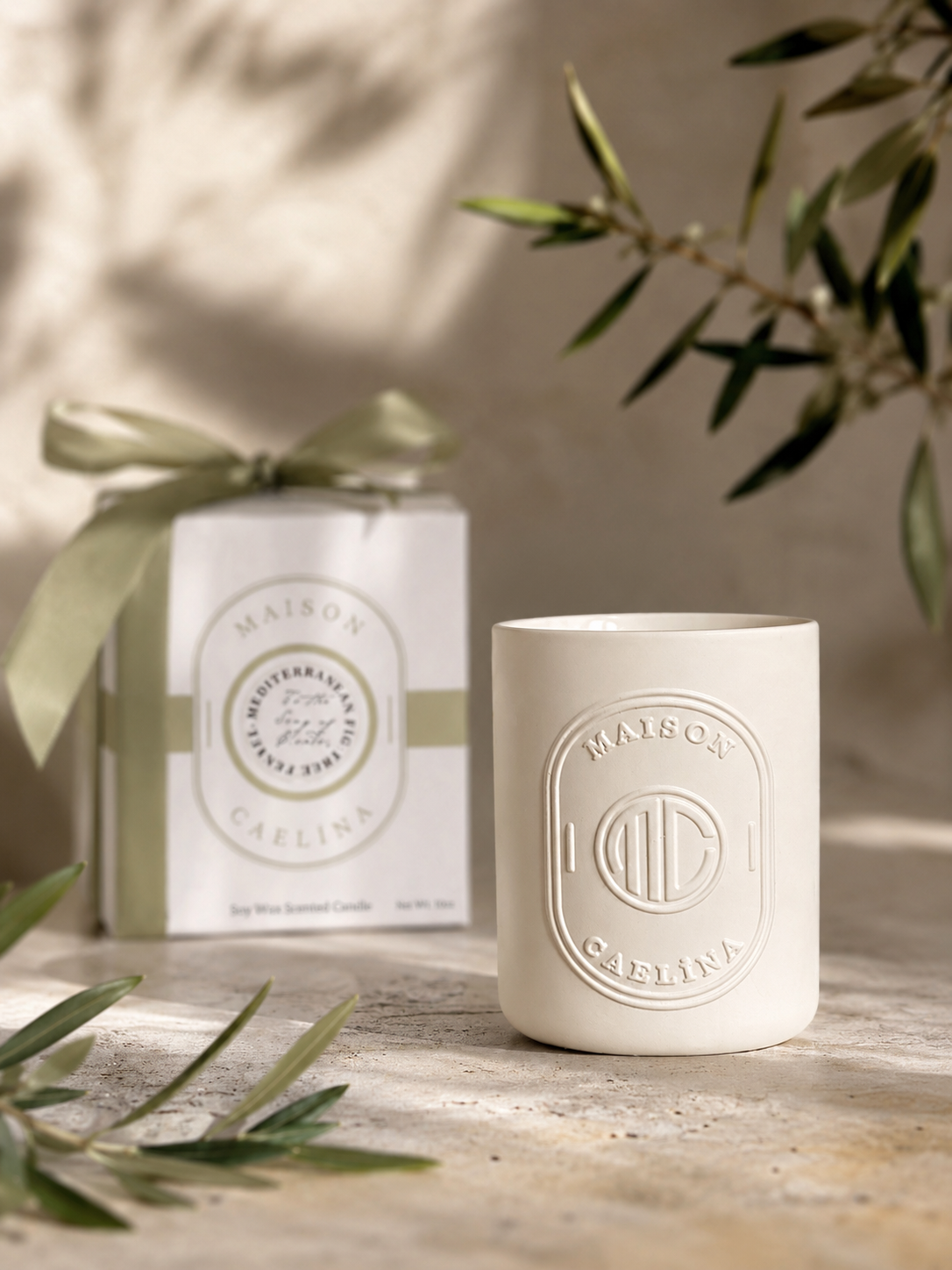 White candle with embossed logo and packaging on a wooden surface with greenery